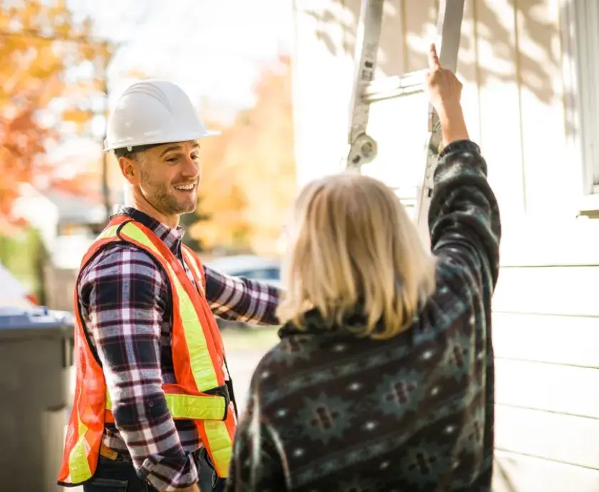 A smiling man wearing a safety helmet and vest smiles while holding a ladder as a woman points to her roof.