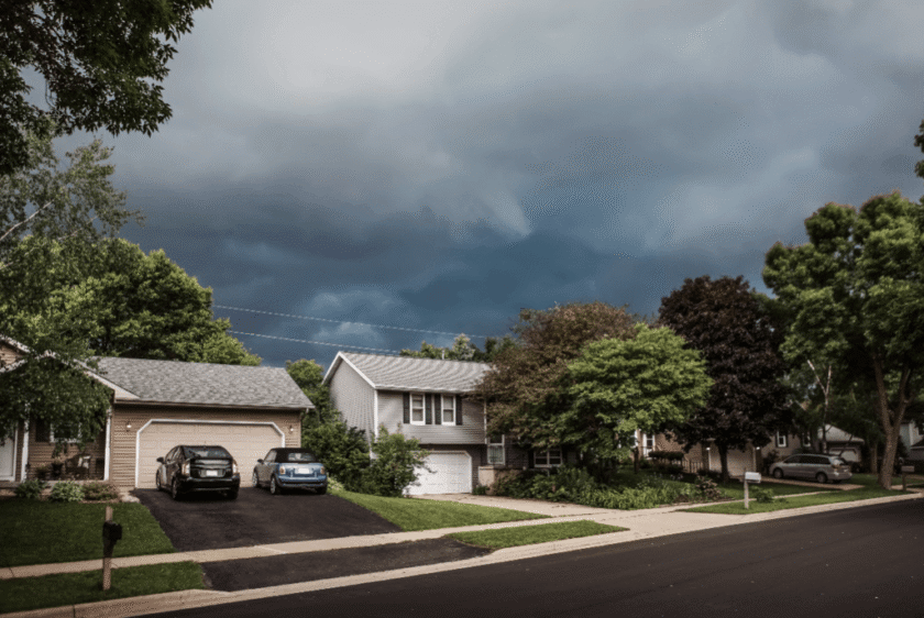 A suburban street with houses, green trees, and parked cars under a dark, cloudy sky, suggesting an approaching storm.