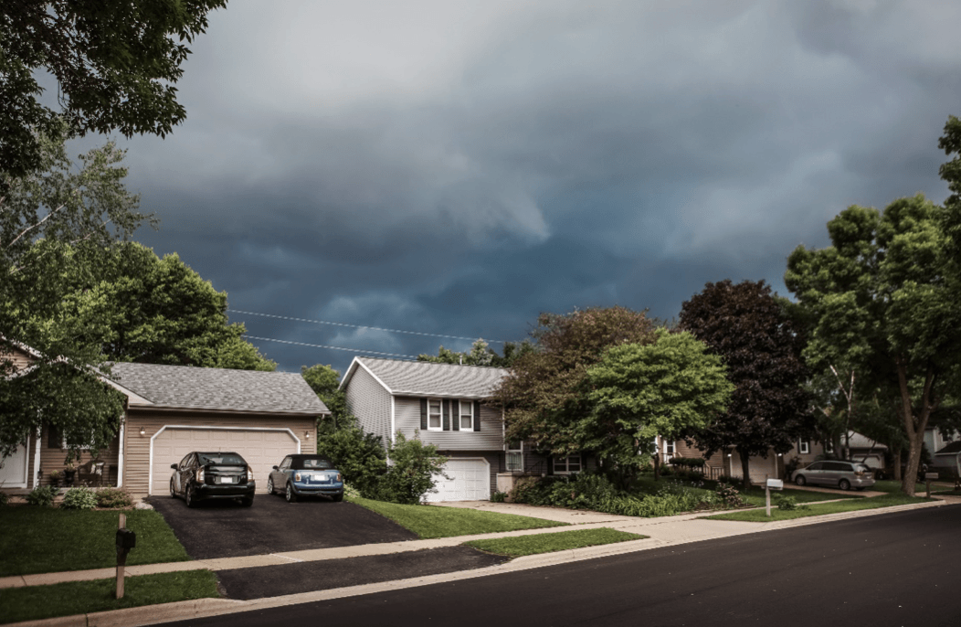 A suburban street with houses, green trees, and parked cars under a dark, cloudy sky, suggesting an approaching storm.