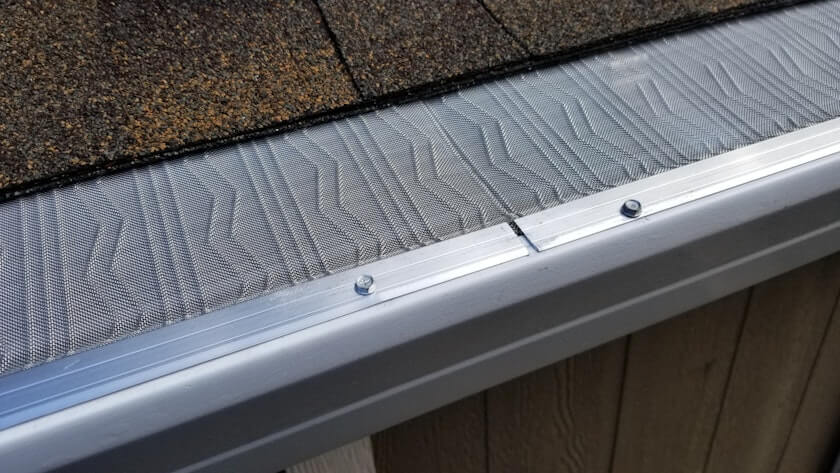 Close-up of a metal gutter guard with a fine mesh cover, securely fastened with screws to the edge of a roof with brown shingles and siding below.