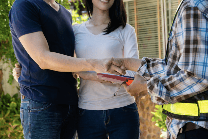A couple stands outside, smiling as the man shakes hands with a worker holding a clipboard. The worker wears a plaid shirt and reflective vest. They appear to be finalizing an agreement or service.